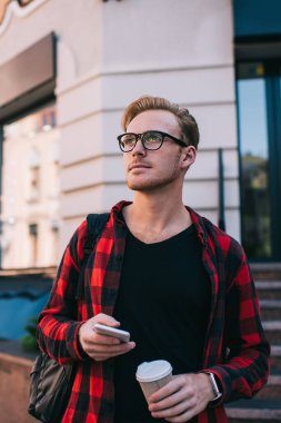 Kendine güveni tam bir erkek. Sırt çantalı ve gözlüklü. Kahve içiyor, sosyal medyayı kontrol ediyor.