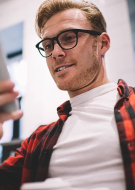 Aşağıdan, gözlüklü barışçıl bir genç adam modern akıllı telefonu dikkatle kullanıyor.