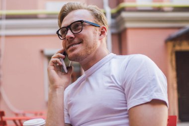 Optik gözlüklü başarılı bir adam halka açık alanda kahve saatinde uluslararası akıllı telefon konuşması yapıyor. Cep telefonu cihazıyla iletişim kuran mutlu bir erkek serbest yazar.