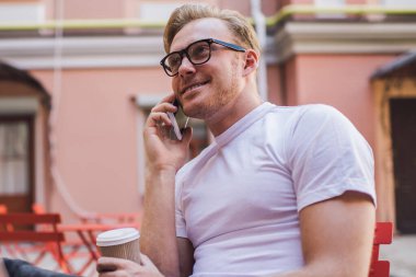 Optik gözlüklü başarılı bir adam halka açık alanda kahve saatinde uluslararası akıllı telefon konuşması yapıyor. Cep telefonu cihazıyla iletişim kuran mutlu bir erkek serbest yazar.