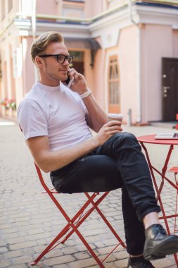 Sokak kafesinde dizüstü bilgisayarla otururken, elinde cep telefonuyla konuşan ve içecek tutan yetişkin bir adamın yan görüntüsü