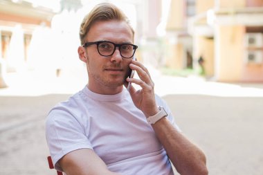 Gözlüklü, kendine güvenen yetişkin bir adam kameraya bakıyor ve bilgisayar kullanırken ve sokak kafesindeki masada otururken akıllı telefonla konuşuyor.