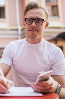 Gözlüklü zeki bir erkek öğrencinin portresi halka açık yerlerde öğrenim sırasında kameraya bakıyor, görüş düzeltmesi için klasik gözlüklü yakışıklı bir adam bilgi defteriyle masada oturuyor.