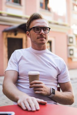 Beyaz tişörtlü düşünceli bir adam sokak kafeteryasında kahve içerken kafasını çeviriyor. Optik gözlüklü şık bir erkek öğrenci elinde kafein içeceğiyle paket kahve tutuyor.