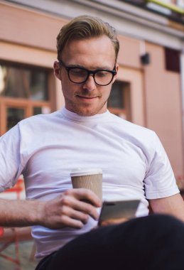 Rahat giyinen, gözlüklü, kahve içen sosyal medyayı kontrol eden, kahve barının önünde cep telefonu kullanan yakışıklı bir genç.