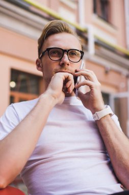 Gözlüklü düşünceli hippi adam uluslararası cep telefonu konuşması yapıyor şehir merkezinde oturuyor ve gezinti bağlantısını kullanıyor. Müşteri hizmetleri ile danışmanlık için tefekkür eden bir adam.