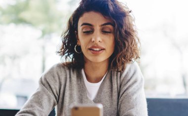 Kısa kıvırcık saçlı, elinde akıllı telefon tutan ve sosyal ağlarda sohbet eden genç bir kadın 4G internet bağlantısı kullanıyor. Hipster kız cep telefonundan blog yazıyor ve web sitesinden haber okuyor.