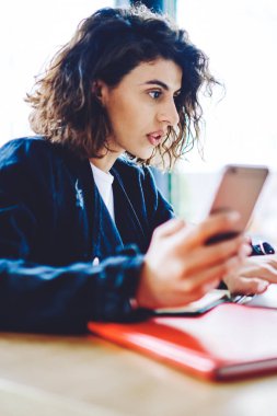 Gündelik giyinmiş kısa kıvırcık saçlı genç bir kadın elinde dijital akıllı telefon tutarken modern çalışma ortamında dizüstü bilgisayarda kablosuz internet kullanarak web sitesinde video izliyor.