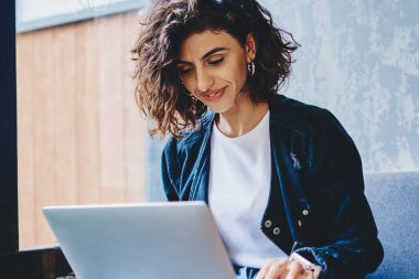 Gülümseyen genç kadın dijital dizüstü bilgisayarda ilginç dersler seyrediyor. Modern mekanda oturan şık hippi kız kablosuz internet bağlantısı kullanarak yazılımı güncelliyor.