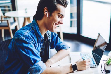 Neşeli erkek öğrenci elektronik kulaklıklarla olumlu müzik listesinden keyif alırken, takipçilere yayın yapmak gibi sosyal ağlarda yayınlanmak üzere ders kitabında eğitim için makale yazıyor.