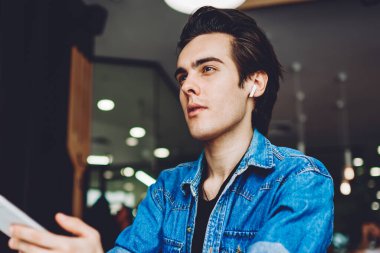 Bluetooth kulaklıklı düşünceli erkek öğrenci, psikolojik ses kitabından gelen bilgiler üzerinde kafa yoruyor, düşünceli hipster adam 4G kablosuz internet ve dinleme kayıtları için cep telefonu uygulaması kullanıyor.