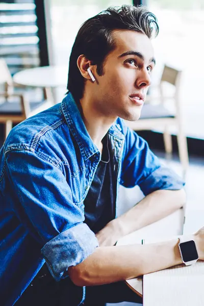 Bluetooth kulaklıklı düşünceli erkek öğrenci, modern teknoloji hakkında e-öğrenim için faydalı tezler yapmak için ders kitabı kullanarak, eğitim sesli kitabından edinilen bilgiler üzerinde yoğunlaştı ve kafa yordu.