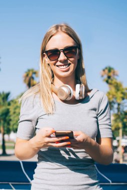 Güneş gözlüklü, kulaklıklı, elinde akıllı telefon olan ve arka planda Barcelona 'nın parlak güneşli manzarasında kameraya gülümseyen mutlu sarışın kadın.