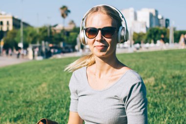 Güneş gözlüklü, kulaklıklı, akıllı telefonlu, yeşil çayır üzerinde otururken kameraya gülümseyen güzel sarışın kadın.