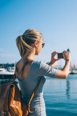 Sırt çantalı sıradan sarışının cep telefonu kullanırken ve şehir parkının parlak güneş ışığı altında fotoğraflarını çekerken, Barcelona, İspanya