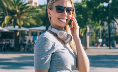 Güneş gözlüklü ve kulaklıklı güzel sarışın kadın güneşli Barcelona şehrinin caddesinde yürüyor ve cep telefonuyla sohbet ediyor.