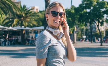 Güneş gözlüklü ve kulaklıklı güzel sarışın kadın güneşli Barcelona şehrinin caddesinde yürüyor ve cep telefonuyla sohbet ediyor.