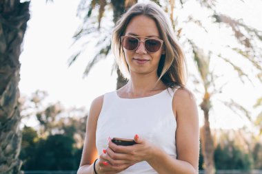Güneş gözlüklü, beyaz yazlık elbise giyen ve Barcelona şehrinde palmiyelerle sokakta duran akıllı telefon kullanan güzel bir kadın.
