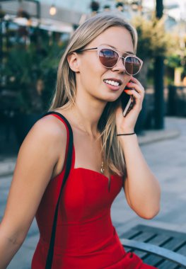 Modern kırmızı üniformalı ve güneş gözlüklü moda sarışın modern şehir binalarına karşı bankta oturuyor ve cep telefonuyla sohbet ediyor.