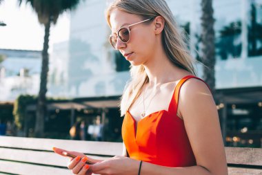 Güneş gözlüklü güzel sarışın Barcelona, İspanya 'nın modern şehir manzarasına karşı güneş ışığında bankta otururken cep telefonu kullanıyor.