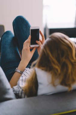 Kadın blogcu, kendi odasındaki rahat kanepeye uzanmış şık bir şekilde sohbet ediyor ve internet siteniz için boş ekran alanı olan modern akıllı telefondan arkadaşlarıyla sohbet ediyor.