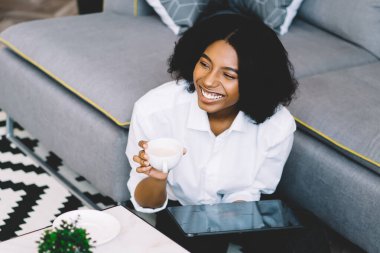 Mutlu siyah kadın kahve molası ve dijital tablet aracılığıyla internet üzerinden iletişim için şık bir halıyla koltuğun yanında dinleniyor, neşeli hippi kız e-postayla gülüyor.