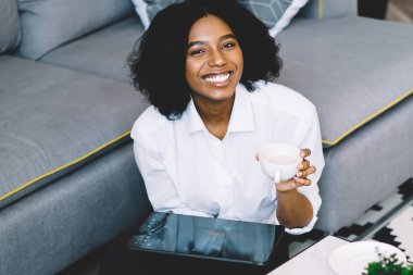 Mutlu Afro-Amerikan hipster kız portresi kahve masasının yanında oturuyor ve boş zamanlarını çay için harcayıp internetten modern dokunmatik ped üzerinden sohbet etmekten zevk alıyor.