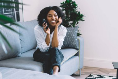 Elinde akıllı telefon zımbırtısıyla gülen koyu tenli hippi blogcu modern dairedeki koltukta otururken şık iç mimari, neşeli Afro-Amerikan genç kadın.