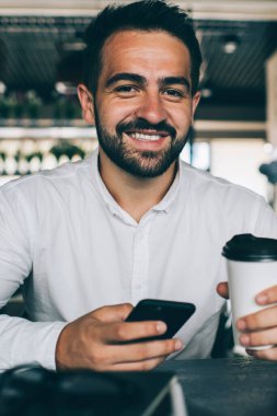 Kahve dükkanının başarılı erkek yöneticisinin portresi elinde cep telefonu cihazıyla internet sitelerini taramak ve dijital ara sırasında kameraya gülümsemek için 4G kablosuz, deneyimli iletişim