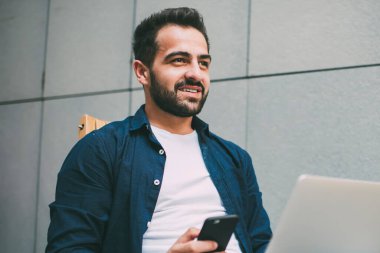 Elinde cep telefonu olan olumlu hipster bir adamın elinde diğer tarafa bakıp gülümserken dizüstü bilgisayar güncellemesi ve serbest çalışma konsepti hakkında danışmanlık için bekleyen programcı bir profesyonelin yan görüntüsü.
