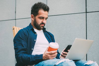 Millennial hipster 'lı adam, modern dizüstü bilgisayarla sokak kafeteryasında akıllı telefondan yetenekli erkek mesajlaşması geliştirmek için otururken internetten cep telefonu uyarısını kontrol ediyor.