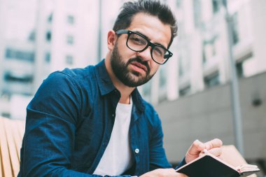 Elinde bilgi kitabıyla kamuya açık alanda kameraya bakan sakallı hippi adamın portresi. Görme koruması için klasik gözlüklü, ciddi bir beyaz adam sınavlara hazırlanıyor.