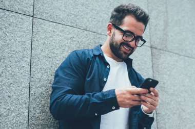 Görme düzeltmesi için optik gözlüklü olumlu erkek blogcu şehir duvarının yanında duruyor ve web sitesinde komik videolar izleyen mutlu hipster adam paylaşım için içerik metni yazıyor