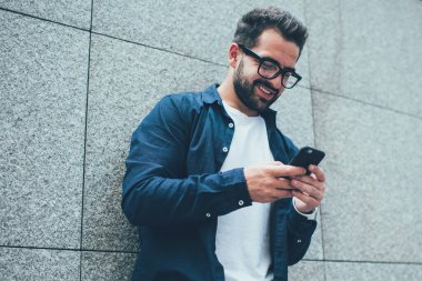 Görme düzeltmesi için optik gözlüklü olumlu erkek blogcu şehir duvarının yanında duruyor ve web sitesinde komik videolar izleyen mutlu hipster adam paylaşım için içerik metni yazıyor