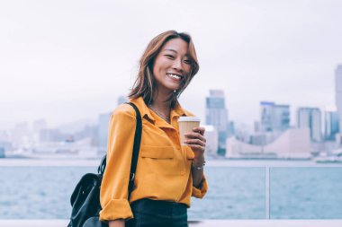 Sırt çantasında tablet ve kahve taşıyan, rüzgarlı bir günde Hong Kong rıhtımında dikilen neşeli, genç bir kadının yan görüntüsü.