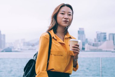 Hong Kong 'da modaya uygun giyinmiş, elinde kahve, elinde dijital tabletle poz veren çekici bir kadın portresi. Nehir kenarındaki şehir merkezinde duran büyüleyici Çinli öğrenci.