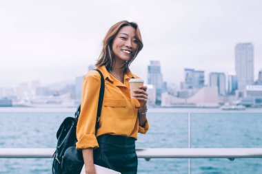 Sırt çantasında tablet ve kahve taşıyan, rüzgarlı bir günde Hong Kong rıhtımında dikilen neşeli, genç bir kadının yan görüntüsü.