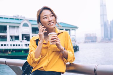 Çekici, neşeli Asyalı kadın, sarı gömlekli, siyah deri etekli, elinde kahve tutan Hong Kong 'daki feribot iskelesinde akıllı telefondan konuşmak için.