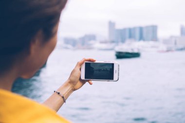 Sarı bluz giyen kadın turistin, çirkin bir günde feribot ve modern gökdelenlerle güzel şehir körfezinin yatay fotoğrafını çekerken görüntüsü.