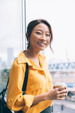 Yarım boy, modaya uygun giyinen, elinde kahve fincanı ve kameraya bakan neşeli bir kadın portresi. Yüzünde sevimli bir gülümseme olan mutlu bir kadın. Çin tatili için boş vakitlerin tadını çıkarıyor.