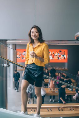 Neşeli Asyalı güzel kadın gündelik giysiler içinde merdivenlerden bir bardak içkiyle çıkarken gülümsüyor ve modern binada kameraya bakıyor.