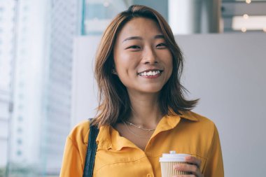 Kafein içmek için boş zamanlarında kameraya gülümseyen neşeli, neşeli, kaygısız Çinli kadının kahveyle gezen neşeli hippi kız portresi.
