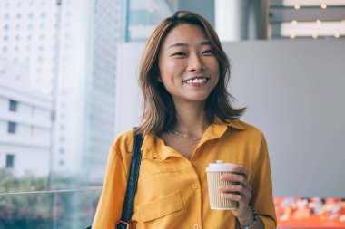 Kafein içmek için boş zamanlarında kameraya gülümseyen neşeli, neşeli, kaygısız Çinli kadının kahveyle gezen neşeli hippi kız portresi.