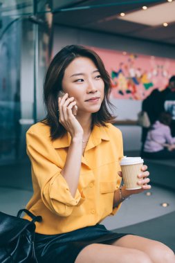 Güzel, sıska, genç Asyalı bir kadının dijital akıllı telefonuyla iletişim kurmasına ve aramasına sevindim. Bir bardak içkiyle birlikte otururken ve modern tasarımla aynı odaya bakarken.