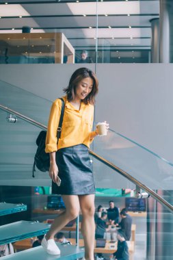 Sarı tişörtlü, gülümseyen Asyalı kadın siyah deri etek ve sırt çantası Hong Kong 'da sıcak içecek ve cep telefonuyla merdivenlerden aşağı iniyor.