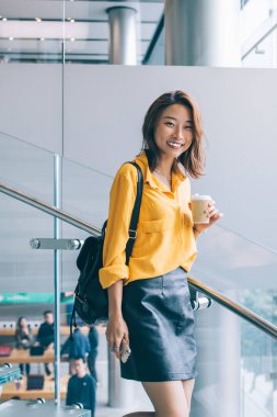 Deri çantalı mutlu Asyalı öğrenci elinde kahve fincanı tutarken ve kameraya gülümserken üniversite arkadaşını, mutluluk konseptini ve neşeli ruh halini bekliyor.