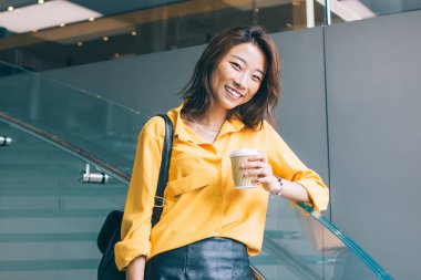 Çekici, mutlu, sarı gömlekli, siyah deri etekli, sırt çantalı, kahve ve cep telefonlu güzel bir Asyalı kadın Hong Kong 'da kapalı kapılar ardında duruyor.