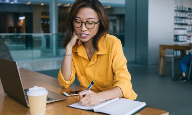 Görüş düzeltmesi için optik gözlüklü çekici Çinli kız öğrenci eğitim için bilgi yazıyor araştırma için zaman harcıyor ve modern dizüstü bilgisayar konseptiyle öğreniyor.