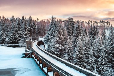 The Laggan Dam (Roybridge Reservoir) and spillway frozen over and covered in snow at sunset clipart