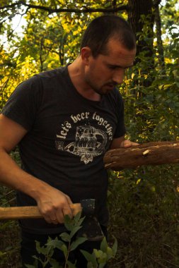 Dalgın bir adam elinde baltayla kütük tutar, o da doğranır. Ormanda akşam vakti erkek portresi.